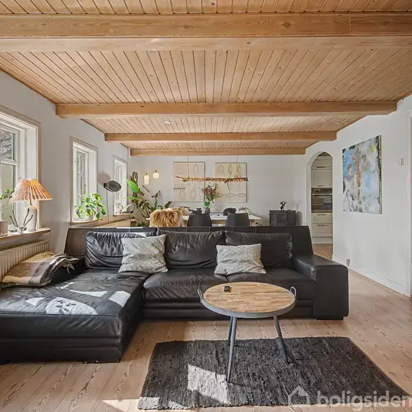 Sort lædersofa står i en stue med trægulv og træloft. Sofaen er placeret ved et vindue med planter. I baggrunden ses et spisebord med blomsterdekorationer.