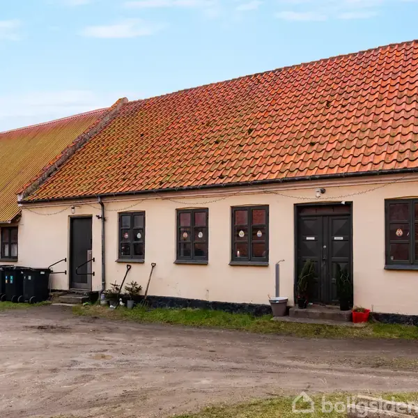Gårdhus med rødt tegltag og beige facade står stille. Papirkurve og planter ved døråbninger. Gruset gårdsplads omgivet af murstensbygninger under blå himmel.