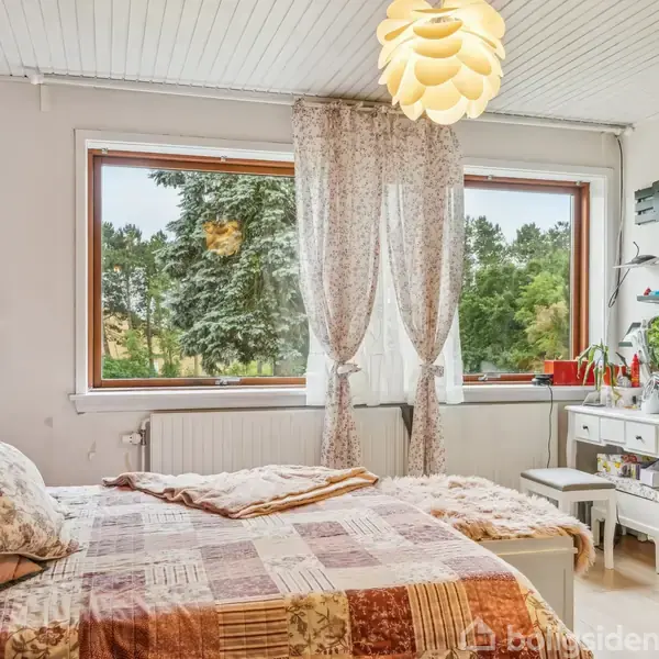En seng med mønstret sengetæppe står ved siden af et stort vindue med gennemsigtige gardiner. Baggrunden viser grønne træer udenfor. Der er et lille bord med stole ved siden af sengen.
