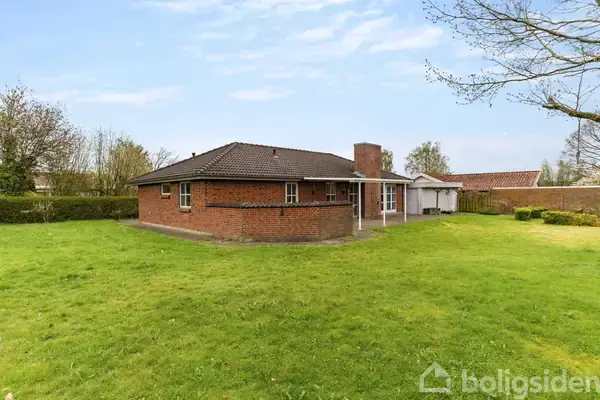 Rødt murstenshus med fladt tag, ingen aktivitet, omgivet af stor græsplæne og træer under blå himmel.