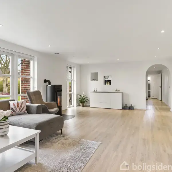 En lys stue med trægulv og store vinduer viser en grå sofa ved siden af et hvidt sofabord med blomster. En brændeovn står ved siden af en skænk, omgivet af minimalistisk indretning.