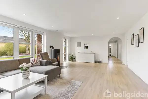 En moderne stue med en grå sofa og hvidt sofabord, står foran store vinduer. En brændeovn står til venstre. Rummet er lyst og rummeligt med trægulv.