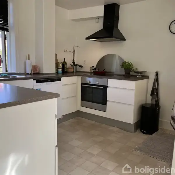 Et moderne køkken med hvide skabe og sort emhætte. Komfuret er midtpunktet, omgivet af køkkenredskaber og planter. Et ur hænger på væggen, og et vindue lader lys ind.