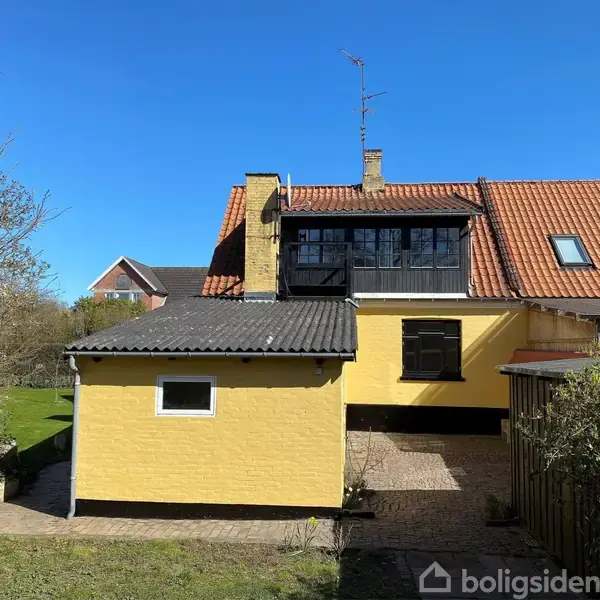 Gult murstenshus med rødt tegltag og en udestue. Ligger i en have omkranset af buske, med klart blå himmel som baggrund.