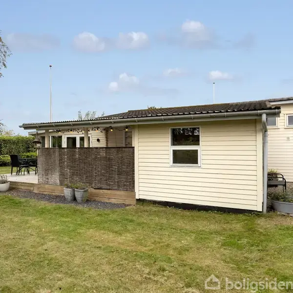 Et lille, lyst træhus står stille med en terrasse og havemøbler i en velplejet have omkranset af hække, under en blå himmel med spredte skyer.