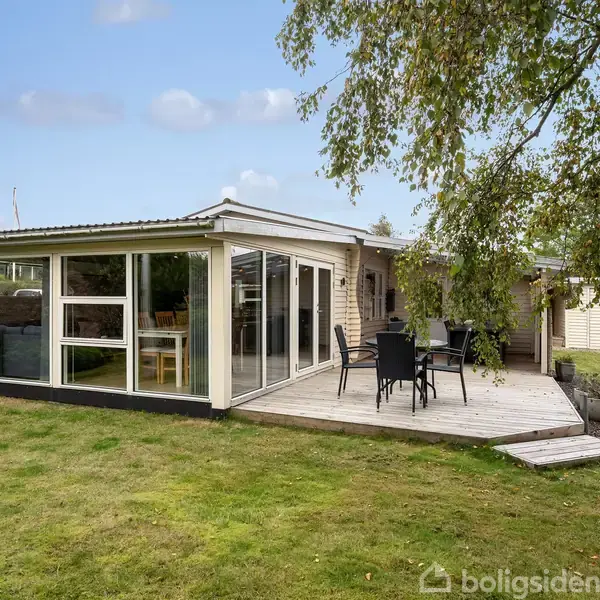 Et lyst sommerhus med store vinduer, der åbner op til en terrasse med havemøbler. Det er omkranset af en grøn græsplæne og træer i baggrunden under en blå himmel.