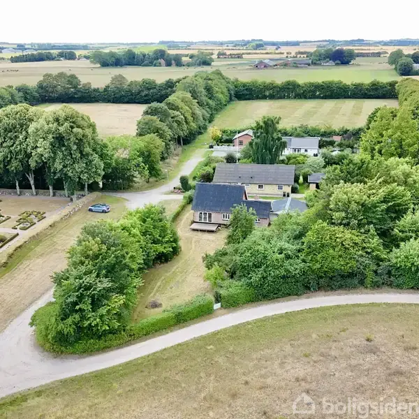 Et hus omgivet af frodige træer og marker nær en lille vej. Ved siden af huset ses velplejede haver og en hvid bygning. Landskabet strækker sig i det fjerne.