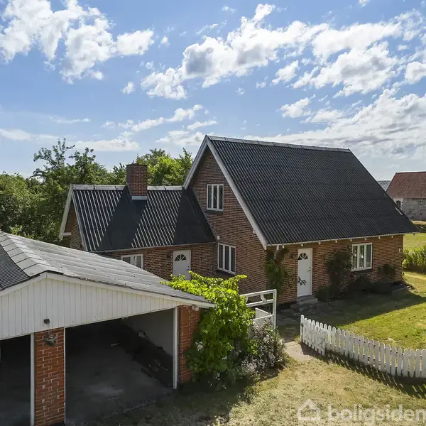 Et rødt murstenshus med sort tag står i en have med grønne buske. Ved siden af huset ses en carport. I baggrunden er der en kirke.