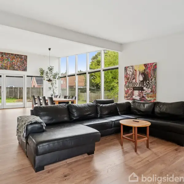 Sort sofa med rundt træbord står i stilfuld og lys stue med store vinduer, betragtelige kunstværker på væggene og trælaminatgulv. En spisestue ses i baggrunden med udsigt til haven.