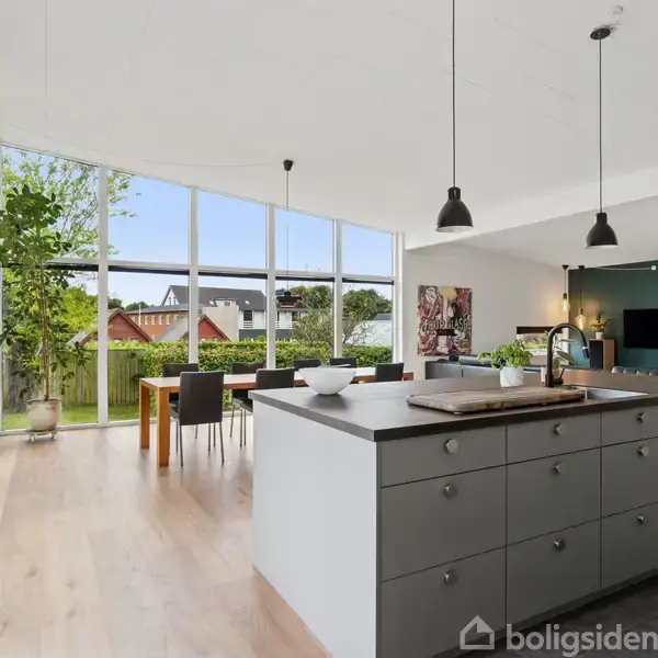 Et moderne køkken med en stor ø, der har skuffer og kogeplade. Spisebordet står i baggrunden ved siden af store vinduer, som giver udsigt til en have.