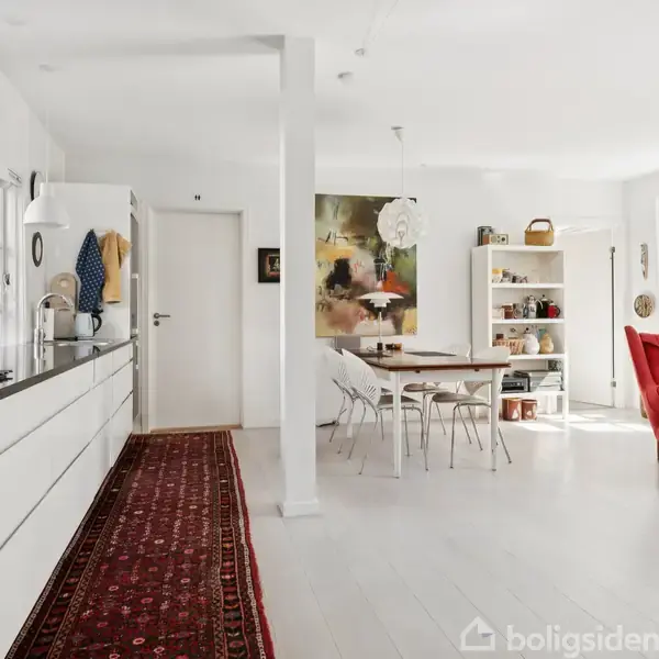 En moderne, lys stue med køkken. Et langt, mørkt tæppe løber langs køkkenet til en spiseplads med hvidt bord og stole. En rød lænestol står ved vinduet. Hylder med bøger og dekorationer i baggrunden.