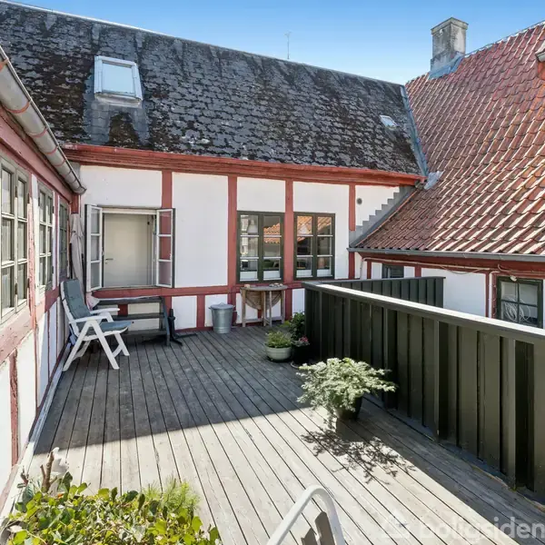 En tagterrasse med trædæk, stole og planter. Det omkringliggende bindingsværkshus har røde teglstenstag og hvidkalkede vægge. Solen skinner, og der er enkelte skyer på himlen.