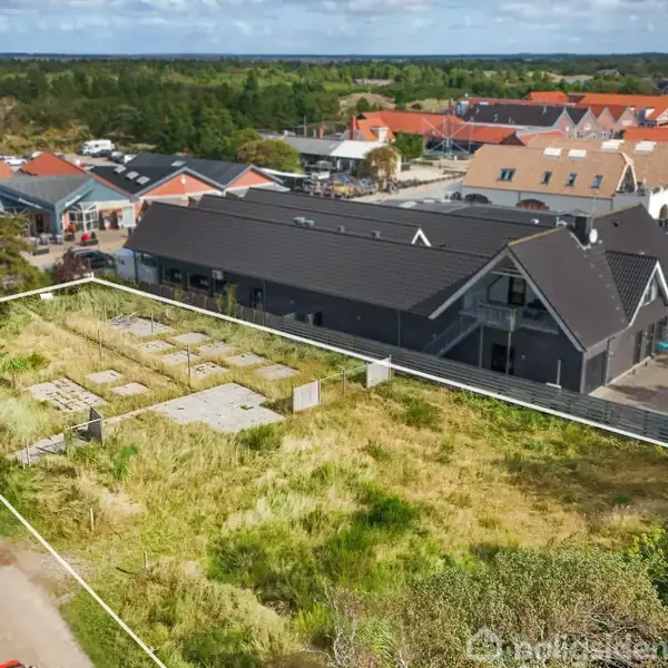 En tom grund med vilde græsser og betonblokke er afgrænset af hvide streger. Ved siden af ligger en stor, moderne bygning i et bymiljø med rødt tag, omgivet af flere huse.