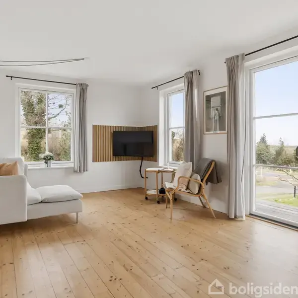 Stue med hvid sofa og træstol, stående mod en hvid væg. Fjernsyn placeret på et lille bord. Store vinduer med gardiner slipper lys ind, omgivet af grøn udsigt.