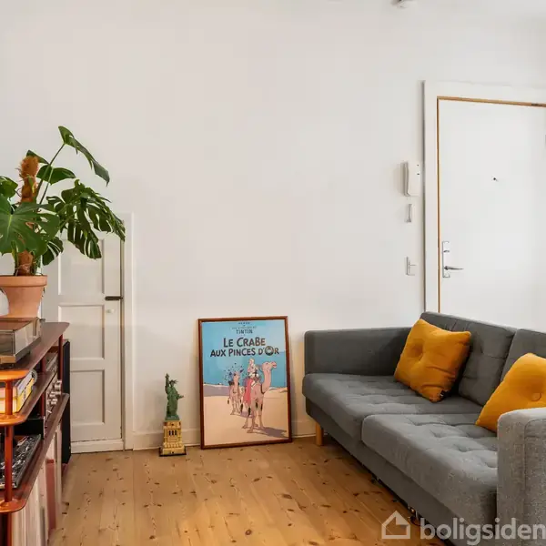 Grå sofa med gule puder står i en stue ved siden af en plakat med teksten "LE CRABE AUX PINCES D'OR," omgivet af en plante og træmøbel. Lyst trægulv og hvid væg.