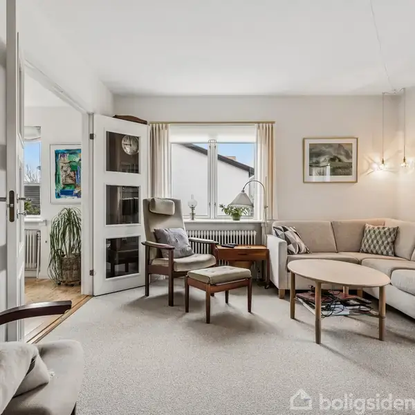 En lys stue med beige sofa og lænestole står omkring et rundt træbord. Væggene har kunstbilleder, og et vindue med lyse gardiner slipper dagslys ind.