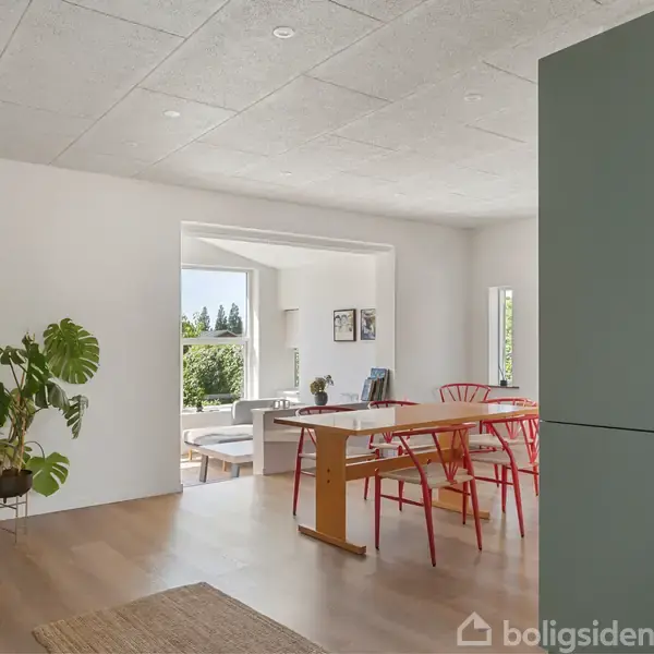 Moderne spisestue med et træbord og røde stole. Store vinduer giver udsigt til grønne træer udenfor. En stor plante og åbne rum skaber en lys og rummelig atmosfære.