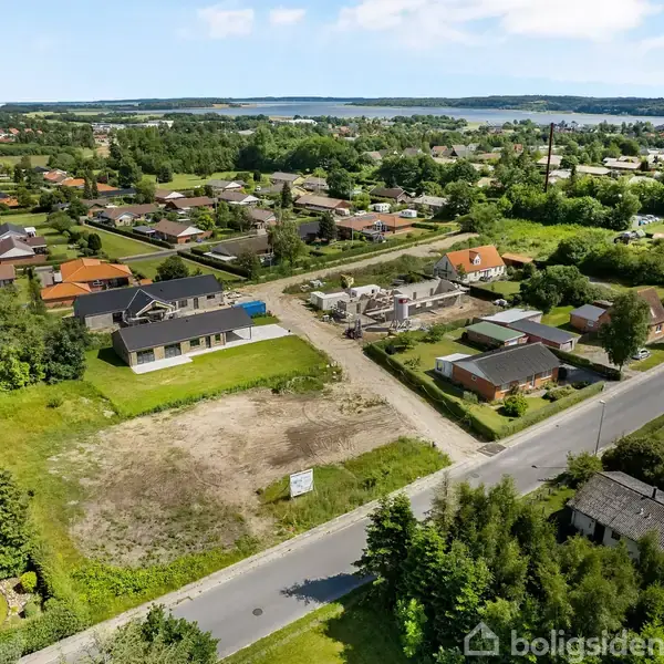 En åben byggegrund ligger ved en villavej i et grønt boligområde nær skove og en fjord. Området er omgivet af huse med forskellige tagformer og haver.