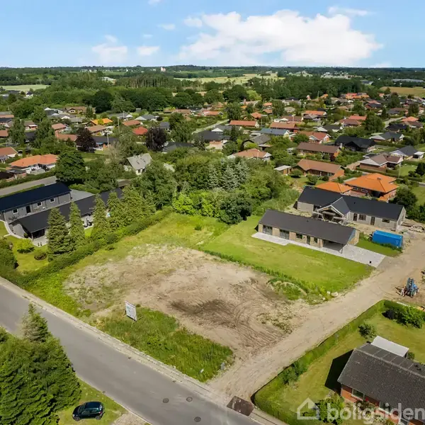 Et ubebygget jordstykke ligger omgivet af en forstad med huse. Grus og ujævnt terræn dominerer grunden, mens grønne haver og veje omgiver det. Boligsiden vandsmærke er synligt.