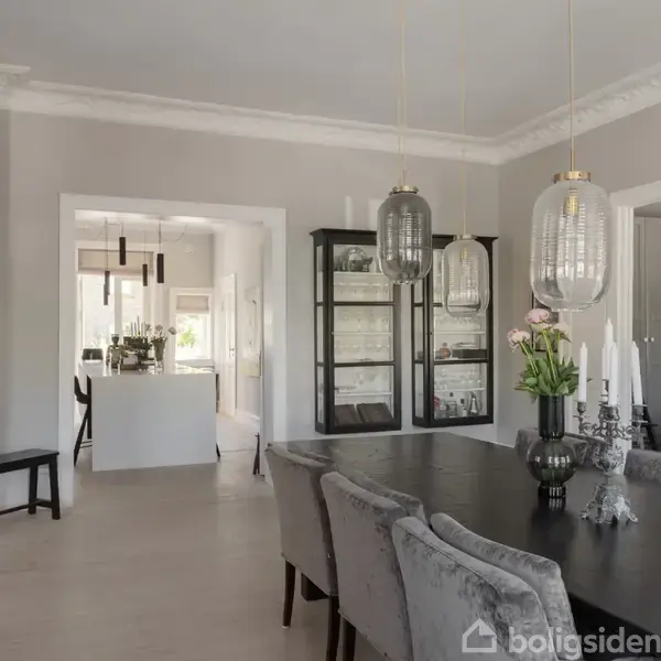 Et elegant spisebord prydes af grå stole og står i et moderne rum med en glasvitrine fyldt med glas. Store, dekorative lamper hænger fra loftet. I baggrunden ses et lyst køkken.