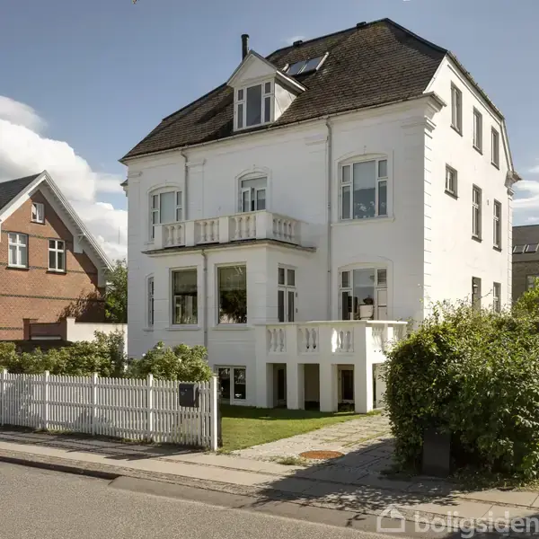 En hvid, tre-etagers villa med skråt tag står ved en gade. Omringet af en hæk og et hvidt stakit, med en velplejet forhave i sollys.