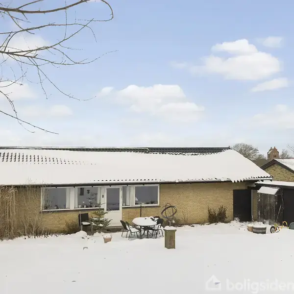 En gul murstensvilla er dækket af sne, omgivet af sneklædte træer og buske under en blå himmel med skyer. Et bord og stole står på terrassen foran huset.