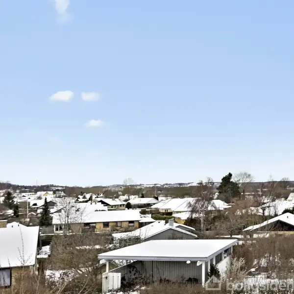 Sneklædte huse står stille i en rolig, vinterlig landsby. Sne dækker tagene i et beboelsesområde, mens en klar, blå himmel strækker sig over det hele.