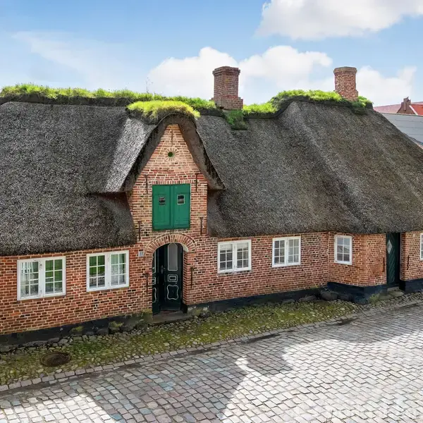 Hus med stråtag og tre skorstensstakke står stille. Bygningen har en murstensfacade med flere hvidrammede vinduer. Det ligger ved en brostensbelagt gade, omgivet af andre bygninger.