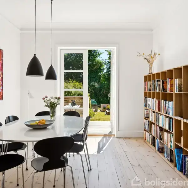 Et stort spisebord med sorte stole står i et lyst spiseområde. Bordet er dekoreret med en skål og vaser. En åben dør fører ud til en have, og en bogreol står i rummet.