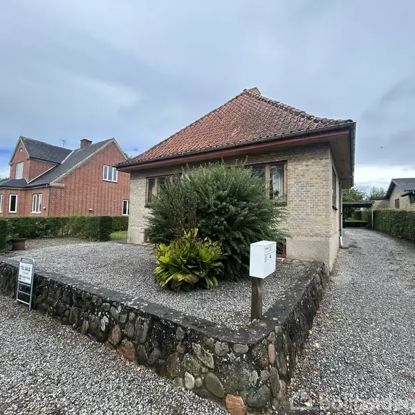 Et gult murstenshus med rødt tegltag står på en grusbelagt grund omkranset af hække. En til salg-skilt ses foran huset, og der er en indkørsel til højre.