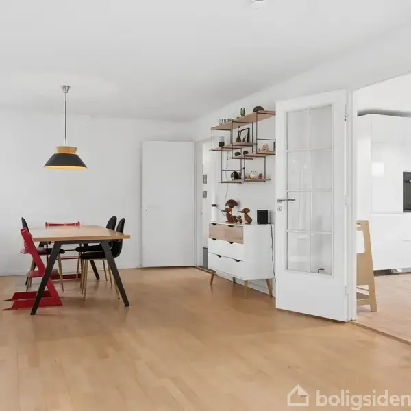 Et spisebord med stole står i et lyst rum med trægulv. Der hænger en lampe over bordet. I baggrunden ses en reol med nips og en åben dør til et andet rum.