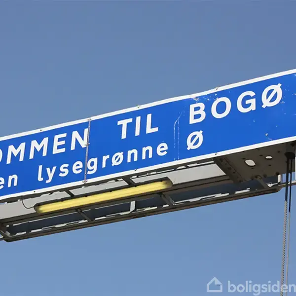 Et blåt vejskilt hænger på en metalstruktur, der viser teksten "VELKOMMEN TIL BOGØ - Den lysegrønne ø" i en klar himmelkontekst.
