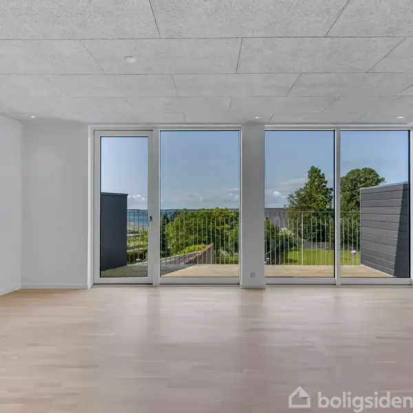 Et lyst, tomt værelse har store gulv-til-loft-vinduer, der giver udsigt til en terrasse og en have med træer. Betongulv og hvidmalede vægge skaber en moderne atmosfære.