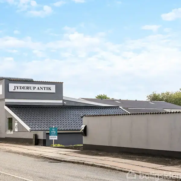 Butikbygning med skiltet "Jyderup Antik" står ved en vej. Bygningen har en lysegrå facade og flere store vinduer. Træer er synlige i baggrunden under en blå himmel.