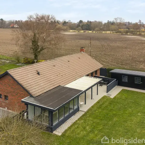 Rødstenshus med brunt tegltag står stille ved siden af en mark i landligt område. Vedhæftede garage og terrasse omkranset af græs og anden bygning i nærheden.