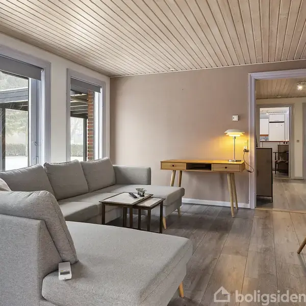 Grå sofa står i en stue med lyst trægulv og træloft. Et skrivebord med lampe er mod væggen. Vinduer til venstre slipper naturligt lys ind.