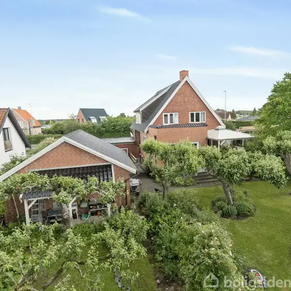 Et rødt murstenshus ses med en grøn have og træer omkransende. Nabohuse med røde tage omgiver det. Huset har en veranda med betagende udsigt over det velholdte område.