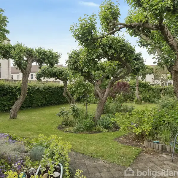 Træer står i en velplejet have med grøn græsplæne. En flisebelagt terrasse har en stol, omkranset af blomsterbede. Baghaven er indrammet af en hæk med huse i baggrunden.