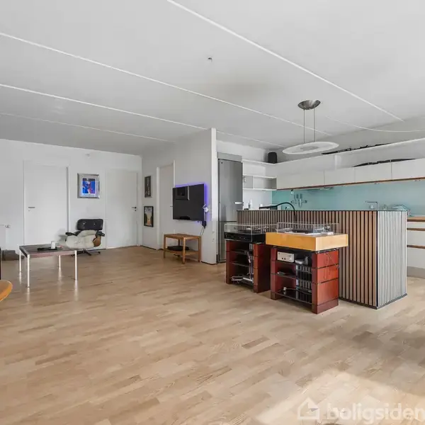 Stue med moderne interiør, åbent køkken med bar, lyse trægulve, og hvidt loft, vægmonteret TV til venstre og stole arrangeret omkring et bord i hjørnet.