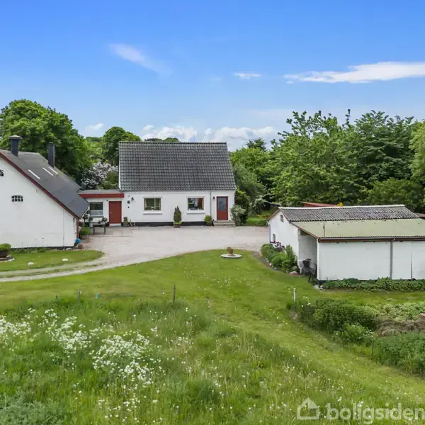 Et hvidt bondehus med gråt tag, omgivet af grønne marker og træer. Der er en grusvej foran huset, som fører til en mindre garagebygning ved siden af.