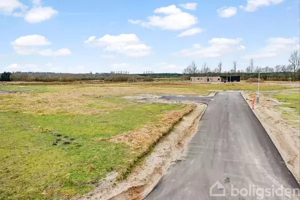 En asfalteret vej strækker sig gennem et åbent område med græs og jord, omgivet af marker og et lavt hus i det fjerne.