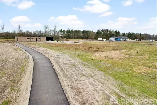 Asfalteret sti fører til minimalistisk bygning under opførelse; omgivet af fladt, åbent græslandskab med spredte bygninger og skov i baggrunden. Klar himmel med få skyer.