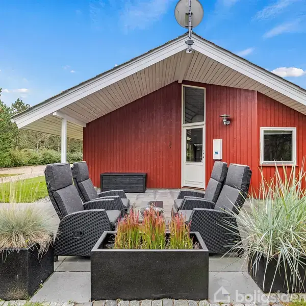 Rødt hus med tagterrasse fyldt med sorte kurvemøbler, står blandt grønne planter og træer. En dør og et vindue ses på huset under en klar, blå himmel.