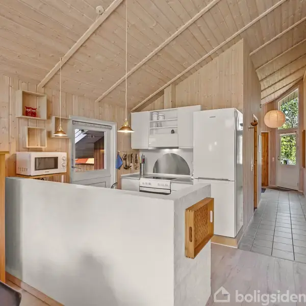 Et køkken med hvidevarer står ved en åben spiseplads; væggene og lofterne er af lyst træ. En gang med flisegulv fører til en dør, og lamper hænger over bordet.