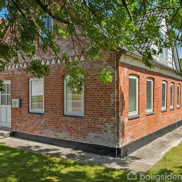 Et rødstenshus står stille omgivet af en grøn græsplæne og skyggefulde træer. Huset har flere hvide vinduer og en hvid hoveddør.