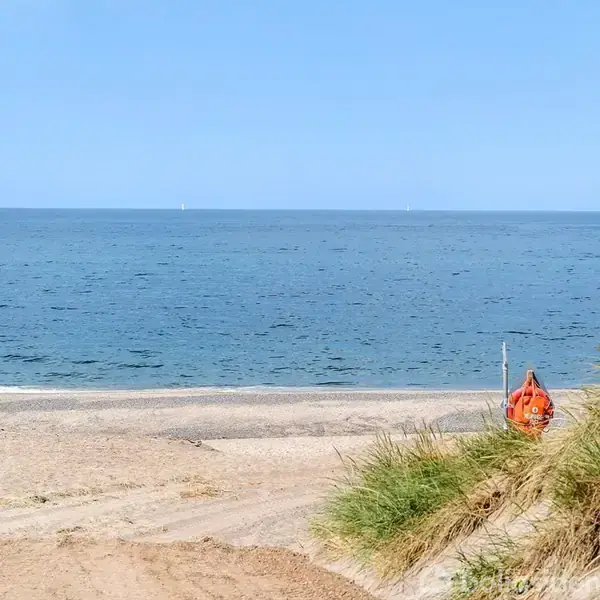 Strand med sand og græsklædte klitter; en orange redningsbøje står på en stang. I baggrunden ses det rolige hav under en blå himmel.