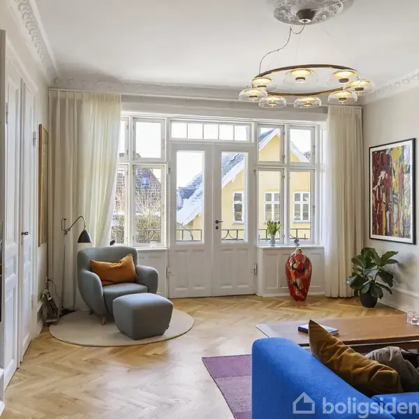 En moderne stue med en blå sofa og lænestol foran lyse vinduer. En dekorativ loftlampe i midten. Kunstværker pryder væggene, mens planter komplimenterer interiøret.