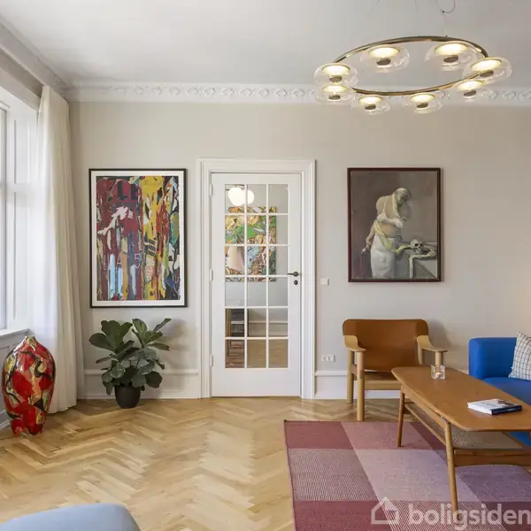 Stue med blå sofa og træbord, omgivet af kunst på væggene. Store vinduer lyser rummet op. Hængelampe i loftet og planter ved siden af døren.
