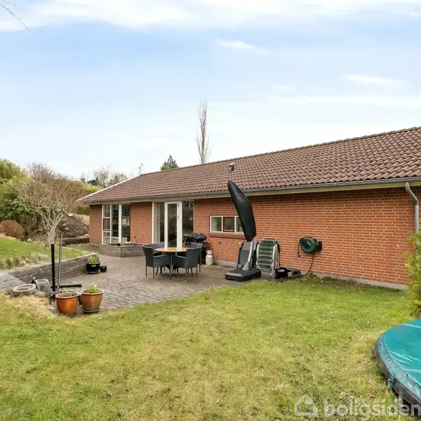 Rødt murstenshus med terrasse, hvor havemøbler og grill står. I forgrunden er en grøn græsplæne med en nedgravet trampolin. Omgivet af buske og træer.