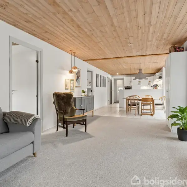 Stue med grå sofa og lænestol, åbner op til et spiseområde med spisebord og stole. Trægulv, træloft, hvide vægge, og en plante ved et klaver. Klar belysning.